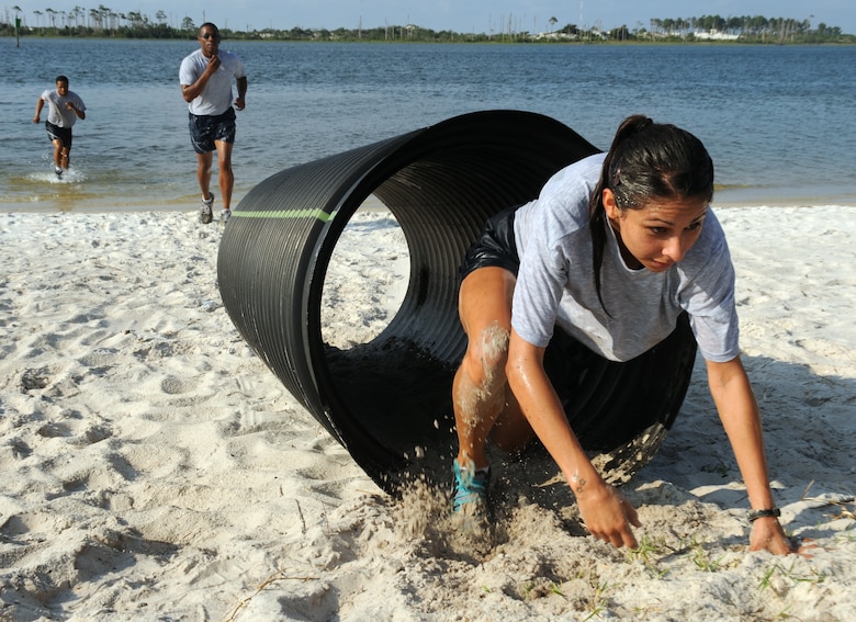 A person emerging from a plastic tunnel obstacle on a beach. Two others running up the shore.